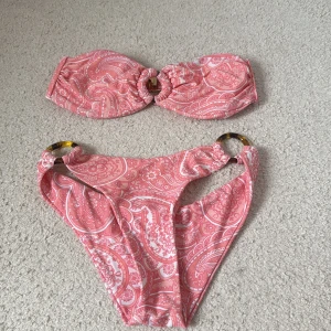 Rosa bandeau bikini med paisleymönster - Säljer en snygg rosa bandeau bikini med paisleymönster och guldfärgade ringdetaljer både på toppen och trosan. Bikinin har en feminin och trendig look, perfekt för stranden eller poolen. Materialet är mjukt och stretchigt syntet för bästa komfort.