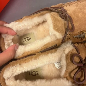 ÄKTA UGG mockasiner med fårskinnsfoder - Mysiga bruna mockasiner från UGG med mjukt vitt fårskinnsfoder. Skorna har dekorativ snörning i mörkbrunt läder och klassisk söm runt tån. Perfekta för kalla dagar när du vill ha det extra bekvämt och varmt om fötterna. Storlek 38 men passar 37 också för det är tofflor. I mycket bra skick men på ena skon är sömmen lite öppen.