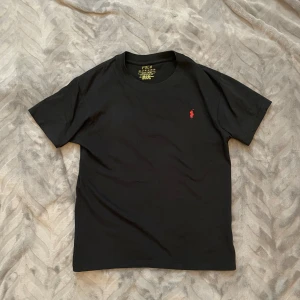 Ralp Lauren T-shirt - Svart T-shirt från Ralph Lauren med en röd broderad logga på bröstet. Skulle säga att den passar allt från 160-170. Tröjan är endast använd ett par gånger men på ett sparsamt sätt. Priset är inte hugget i sten och skriv gärna vid minsta fundering.