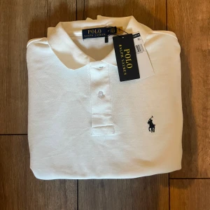 Vit pikétröja från Polo Ralph Lauren - Vit pikétröja från Polo Ralph Lauren med klassisk krage och två knappar framtill. Tröjan har det ikoniska broderade polospelar-loggan i marinblått på bröstet. Tillverkad i mjuk bomull som andas och passar perfekt till en clean och stilren look.