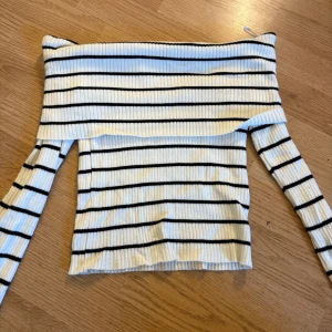 Randig offshoulder topp från H&M - Säljer en ribbad offshoulder topp från H&M i vitt med svarta horisontella ränder. Toppen har lång ärm och är figurnära med en snygg croppad passform. Perfekt för dig som gillar en stilren och trendig look.