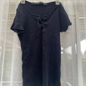 Svart ribbad topp med snörning - Svart ribbad topp från The Black Label Collection med korta ärmar och snygg snörning framtill vid halsen. Mjuk och stretchig bomullskänsla, perfekt till jeans eller kjol. Enkel men ändå med en cool detalj framtill.