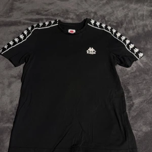 Svart Kappa t-shirt med logoband - Svart t-shirt från Kappa i bomull med klassiskt logoband i vitt längs axlarna och ärmarna. Kappa-logga tryckt på bröstet. Enkel och sportig design som passar perfekt till streetwear eller chill vardagsstil. Jag köpte en t-shirt på herravdelningen, men den passar som unisex. Jag är en tjej som är 170-172 cm lång.