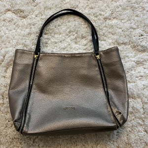 Guess metallic axelväska med dragkedjor - Snygg metallicfärgad axelväska från Guess med svarta handtag och gulddetaljer. Väskan har två dekorativa dragkedjor framtill och magnetlås. Invändigt finns fack och svart foder, väskan är rymlig. Perfekt för dig som gillar stilrena accessoarer med lite edge. Defekter förekommer så varan är sliten på utsidan som syns på bilden.🩷
