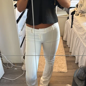 Vita tighta byxor med söm framtill - Säljer ett par vita byxor med låg midja och tight passform. Byxorna har dekorativa sömmar framtill och två knappar i midjan. Materialet är stretchigt och bekvämt, perfekt för en clean och fräsch look. Har 2 sömmar som gått upp går att laga