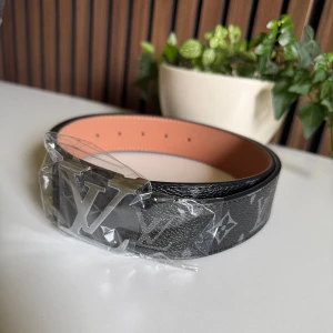 Louis Vuitton svart skärp med monogram (9) - Säljer ett Louis Vuitton bälte i svart med klassiskt monogrammönster och stor LV-spänne i silver. Bältet är tillverkat i exklusivt skinn med beige insida och har en bred, stilren form som ger en lyxig vibe. Perfekt för dig som vill ha en statement-accessoar.