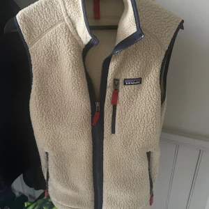 Beige fleeceväst från Patagonia (S) - Säljer en beige fleeceväst från Patagonia i storlek S. Västen har hög krage, dragkedja framtill och flera fickor med röda detaljer på dragkedjorna. Perfekt för lager-på-lager och outdoor-vibes. Mjuk och fluffig känsla, klassisk Patagonia-logga på bröstet. Riktigt fet väst som sagt passar till allt. 