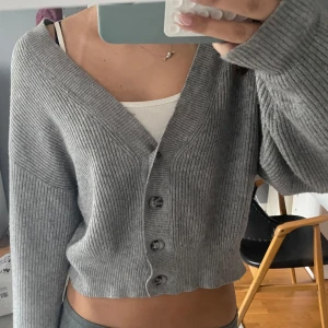 Grå croppad kofta med knappar - Säljer en grå ribbstickad kofta med croppad passform och stora knappar framtill. Koftan har djup v-ringning och långa ärmar, perfekt att slänga över ett linne. Supermjuk och enkel att styla till olika outfits.
