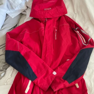 Röd vindjacka från Tenson - Röd vindjacka från Tenson med svarta detaljer på ärmarna och vita dragkedjor. Jackan har huva, långa ärmar och praktiska fickor. Perfekt för blåsiga dagar och outdoor-äventyr. Snygg och sportig look med tydliga loggor på bröstet och ärmen. storlek L, ny kostar runt 1000kr