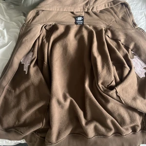 Brun Sweet SKTBS hoodie med dragkedja - Säljer en brun hoodie från Sweet SKTBS med broderad text på bröstet i vit och svart. Tröjan har huva, dragkedja och mjukt fleecefoder på insidan. Perfekt för dig som gillar streetwear och vill ha något bekvämt och stilrent.