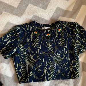 Mönstrad croppad topp från Zara - Säljer en croppad topp från Zara med blå bas och limegröna bladmönster. Toppen har korta ärmar och är dekorerad med färgglada pärl- och stenapplikationer vid halsen. Materialet är stretchigt och strukturerat, perfekt för dig som gillar unika detaljer.