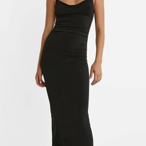 Svart långklänning med slits - Supersnygg svart långklänning med smala axelband och draperad urringning. Klänningen har en tight passform och en slits bak för extra edge. Materialet är mjukt och stretchigt, vilket gör den bekväm att bära hela kvällen. Perfekt för dig som gillar minimalistisk stil med en twist.
