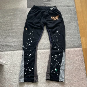 Gallery Dept. svarta mjukisbyxor M - Svarta mjukisbyxor från Gallery Dept. med elastisk midja, färgstänk och tryckt logga på ena benet. Byxorna har grå och mörkgrå paneler längs sidorna och är tillverkade i 100% bomull. Snygg streetstil med unik färgdetalj.