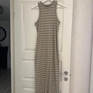 Beige och vit randig långklänning - Snygg långklänning i beige med vita horisontella ränder. Ärmlös modell med rund halsringning och ribbad struktur. Perfekt för en avslappnad och trendig look under varma dagar.