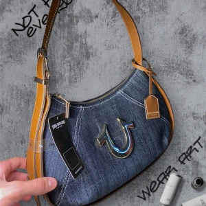 True Religion jeans handväska / axelväska  - Snygg axelväska från True Religion i blått jeanstyg med bruna skinn-detaljer och guldfärgad logga framtill. Väskan har dragkedja upptill och ett ljust innerfoder med mönster samt innerfack. Perfekt för dig som gillar street och denim vibes. Helt ny!!!