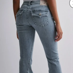 True Religion Joey Low Rise Flare Jeans - Säljer ett par ljusblå True Religion Joey jeans med låg midja och utsvängda ben. Klassiska bakfickor med lock och silverknappar, samt tydliga sömmar. Jeansen är i mjuk denim 