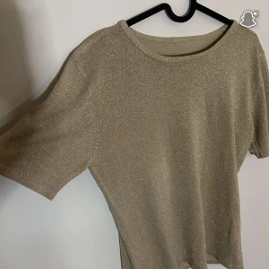 Glittrig guld topp med kort ärm - Snygg guldig topp med glittrigt tyg och korta ärmar. Perfekt för dig som vill sticka ut lite extra med en enkel men ändå festlig look. Rund halsringning och mjukt material som känns skönt mot huden.