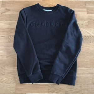 Svart sweatshirt från Calvin Klein Jeans - Svart sweatshirt från Calvin Klein Jeans med broderad logga framtill. Klassisk rund halsringning och långa ärmar. Mjuk insida och ribbade muddar vid ärmslut och nederkant. Perfekt för en clean och stilren look.