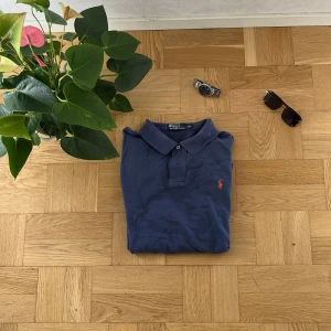 Blå långärmad pikétröja från Polo Ralph Lauren - Klassisk blå långärmad pikétröja från Polo Ralph Lauren med orange broderad logga på bröstet. Tröjan har krage, knappar vid halsen och är tillverkad i mjuk bomull. Perfekt för dig som gillar en avslappnad men ändå stilren look.