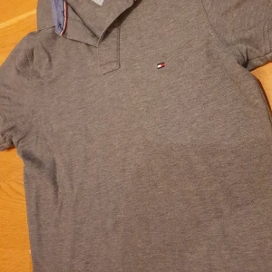 Grå pikétröja Tommy Hilfiger S - Äkta T-Shirt som är snygg grå pikétröja från Tommy Hilfiger i slim fit-model/164-170. Klassisk krage med diskret logga på bröstet och randigt tyg på insidan av kragen. Tillverkad i bomull och perfekt för en clean och stilren look.