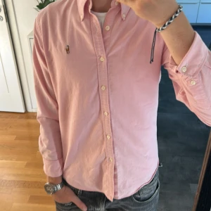 Rosa skjorta från Ralph Lauren - Snygg rosa skjorta från Ralph Lauren i slim fit-modell. Skjortan har klassisk krage, knäppning framtill och broderad logga på bröstet. Tillverkad i 100% bomull och har långa ärmar med knappar vid manschetterna. Perfekt för en clean och stilren look. Priset kan såklart diskuteras 