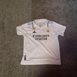 Real Madrid Benzema matchtröja + shorts - Säljer en vit Real Madrid matchtröja med Benzema och nummer 9 på ryggen, samt tillhörande shorts. Tröjan har korta ärmar, krage och broderade klubbmärken. Adidas-logga och lila detaljer på axlar och shorts. Materialet är lätt och andas, perfekt för sport personer eller för skolan.