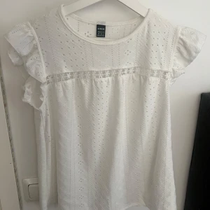 Vit blus med spetsdetaljer från SHEIN - Söt vit blus från SHEIN med broderade hålmönster och volangärmar. Blusen har en fin spetsdetalj med små blommor över bröstet och rund halsringning. Perfekt för dig som gillar romantisk och feminin stil.