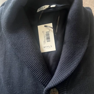 Mörkblå kofta från RYVLS Stockholm - Snygg mörkblå kofta från RYVLS Stockholm med bred krage och knappstängning framtill. Koftan är stickad i mjukt material och har en klassisk, stilren look som passar perfekt till jeans eller chinos. Perfekt för dig som gillar enkel och clean stil.
