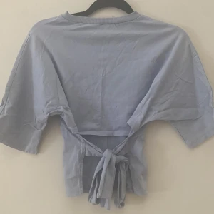 Ljusblå knyt-topp från Nelly - Supersöt ljusblå topp från Nelly i bomull med korta ärmar och snygg knytning i ryggen. Perfekt för dig som gillar en enkel men trendig look. Rund halsringning och loose fit gör den extra bekväm. Säljer billigt så den har en liten fläck på sig 