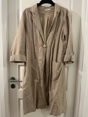 Beige linnékappa från Gina Tricot - Säljer en beige kappa från Gina Tricot i linnematerial. Kappan har en avslappnad passform, stora fickor framtill, slag vid kragen och en enkel knappstängning. Perfekt för dig som gillar en stilren och enkel look med uppvikta ärmar.