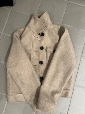 Beige overshirt med stora knappar - Snygg beige overshirt i ullmix med stora svarta knappar framtill och två bröstfickor med lock. Rak passform och hög krage ger en clean och modern look. Perfekt att slänga över en hoodie eller t-shirt för extra lager.
