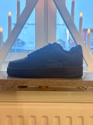 Nike Air Force 1 helsvarta sneakers - Säljer ett par klassiska Nike Air Force 1 i helsvart skinn. Skorna har perforerad tå, rund tå, snörning och en chunky platt sula. Perfekta för dig som vill ha en clean och tidlös look med streetkänsla. AF1-detalj på snörningen och diskret Nike-logga på sidan.