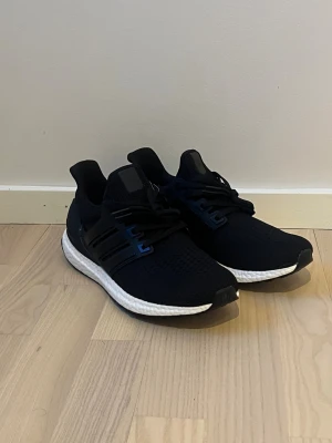 Svarta Adidas Ultraboost sneakers - Säljer ett par svarta Adidas Ultraboost sneakers med vit sula och svart ovandel i mesh. Skorna har klassiska tre ränder på sidan, snörning och en bekväm, responsiv sula som passar perfekt för både träning och vardag. Stilrena och sportiga med modern design.