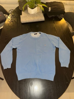 Ljusblå stickad tröja från Gant - Säljer en ljusblå stickad tröja från Gant med rund halsringning och diskret broderad logga på bröstet. Tröjan har lång ärm och ribbade muddar vid ärmslut och nederkant. Perfekt för dig som gillar klassisk och stilren design.