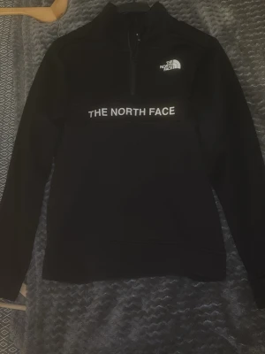 Svart half zip tröja The North Face - Säljer en svart långärmad tröja från The North Face med vit logga på bröstet och stor text över magen. Tröjan har half zip-dragkedja och hög krage, perfekt för lager på lager. Materialet känns mjukt och sportigt. Storleken är XL i barnstorlek vilket går att jämföra med XS i vanliga storlekar.