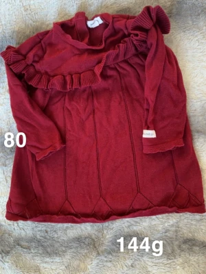 Vinröd klänning med volang från Newbie - Supersöt röd klänning från Newbie med långa ärmar och volangdetalj över bröstet.  perfekt för mysiga dagar. Snygga sömmar nertill och en klassisk rund halsringning.🌸Fint använt skick. Storlek 80