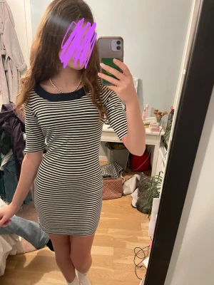 Randig kortklänning med marinblå krage - Säljer en söt kortklänning med smala svarta och vita ränder och en marinblå krage. Klänningen har korta ärmar och en figurnära passform som framhäver siluetten. Perfekt för dig som gillar en enkel men stilren look.