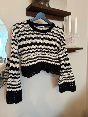 Svartvit stickad croppad tröja Zara - Säljer en croppad stickad tröja från Zara i svart och vitt med vågigt randigt mönster. Tröjan har vida långärmar och är virkad med hålmönster för en luftig känsla. Perfekt för dig som gillar en trendig och unik look.
