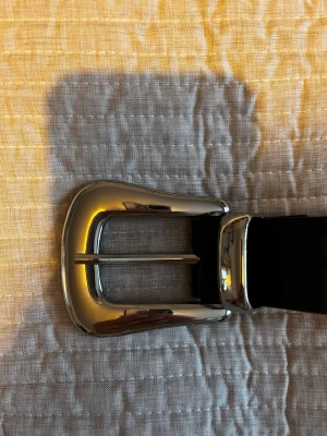 Svart bälte från Gina Tricot - Stilrent svart läderskärp med blank silverfärgad spänne i klassisk design. Passar både till vardag och mer uppklädda tillfällen.  Det finns en mindre defekt i lädret som syns på sista bilden, men den påverkar inte funktionen. Ett snyggt och prisvärt skärp i bra begagnat skick.