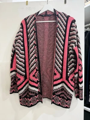 Mönstrad kofta i rosa och svart - Säljer en stickad kofta från New Yorker med grafiskt mönster i rosa, svart och vitt. Koftan är öppen framtill, har lång ärm och är perfekt för lager-på-lager. Den har en loose passform och coola detaljer i mönstret.