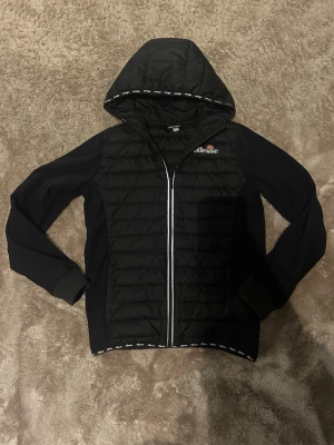 Svart pufferjacka från Ellesse - Säljer en svart pufferjacka från Ellesse med huva och hel dragkedja framtill. Jackan har quiltad front, mjuka muddar vid ärmsluten och logga på bröstet. Snygga detaljer med Ellesse-tryck längs huva och nederkant. Perfekt för kyliga dagar.