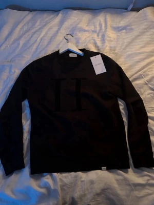 Svart tröja från Les Deux - Säljer en svart sweatshirt från Les Deux med stor, diskret logga framtill. Tröjan har rund halsringning och långa ärmar. Enkel och stilren design som passar perfekt till jeans eller chinos.