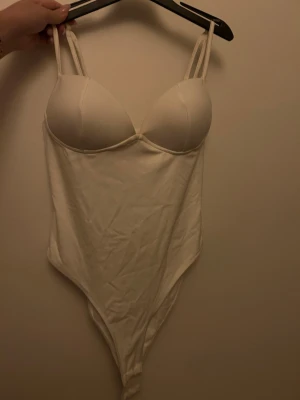Vit korsettinspirerad bodysuit - Säljer en vit korsettinspirerad bodysuit med vadderade kupor och dubbla smala axelband. Toppen har en clean och enkel design med mjukt stretchigt material som formar sig efter kroppen. Perfekt att styla med jeans eller kjol för en trendig look.