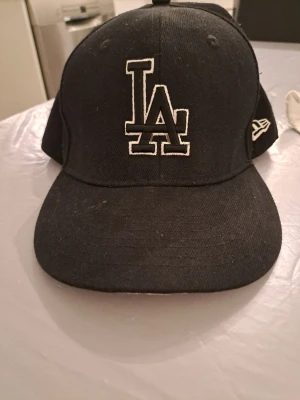 Svart LA New Era 59FIFTY keps - Säljer en svart New Era 59FIFTY keps från Cooperstown Collection med LA-logga broderad i vitt framtill och MLB-logga baktill. Klassisk form med böjd skärm och broderade ventilationshål. Perfekt för dig som gillar streetwear och sport.