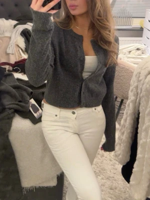 Mörkgrå stickad kofta med knappar - Mysig mörkgrå stickad kofta med rund hals och knäppning framtill. Koftan har lång ärm och en lite loose passform, perfekt att slänga över ett linne eller t-shirt. Enkel och stilren design som passar till det mesta.