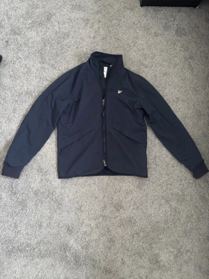Overshirt/jacka från Lyle and Scott - Xs men sitter som S! Modellen är 180 och som man ser så är det lite liten för modellen! Nypris 1500kr! Säljer en marinblå jacka från Lyle & Scott med dragkedja och två snedställda fickor framtill. Jackan har ribbade muddar vid ärmsluten och en liten broderad logga på bröstet. Perfekt till vår och höst, enkel och stilren design.