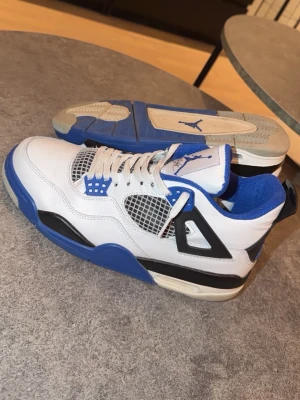 Nike Air Jordan 4 Retro blå/vit - Nike Air Jordan 4 Retro sneakers i vit och blå med svarta detaljer. Skorna har meshpaneler på sidorna, snörning och Jumpman-logga på plösen och hälen. Tillverkade i skinn och syntetmaterial med platt sula och rund tå. Perfekt för dig som gillar klassiska och sportiga sneakers.