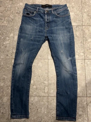 John Richmond jeans med slitningar - Säljer ett par blå slim fit jeans med slitna detaljer och klassisk femficksdesign. Jeansen har en snygg tvättad look, normalhög midja och svart JR-logga på myntfickan. Perfekta till sneakers och hoodie.