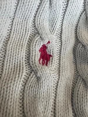 Vit kabelstickad kofta från Polo Ralph Lauren - Vit kabelstickad kofta från Polo Ralph Lauren i 100% bomull. Klassisk v-ringning, knappar framtill och en liten rosa broderad logga på bröstet. Snygg ribbad kant nertill och vid ärmslut. Perfekt för lager-på-lager under hösten eller våren.