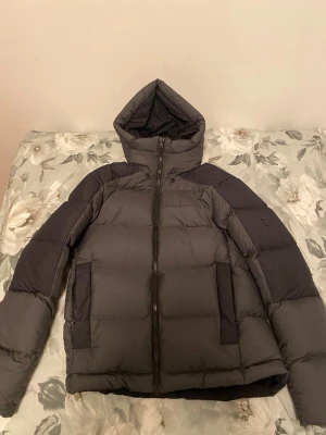 Mörkgrå Peak Performance pufferjacka - Säljer en mörkgrå pufferjacka från Peak Performance med huva och dragkedja framtill. Jackan har två stora fickor, diskret logga på bröstet och en snygg detalj på ärmen. Perfekt för kalla dagar och riktigt skön att bära.